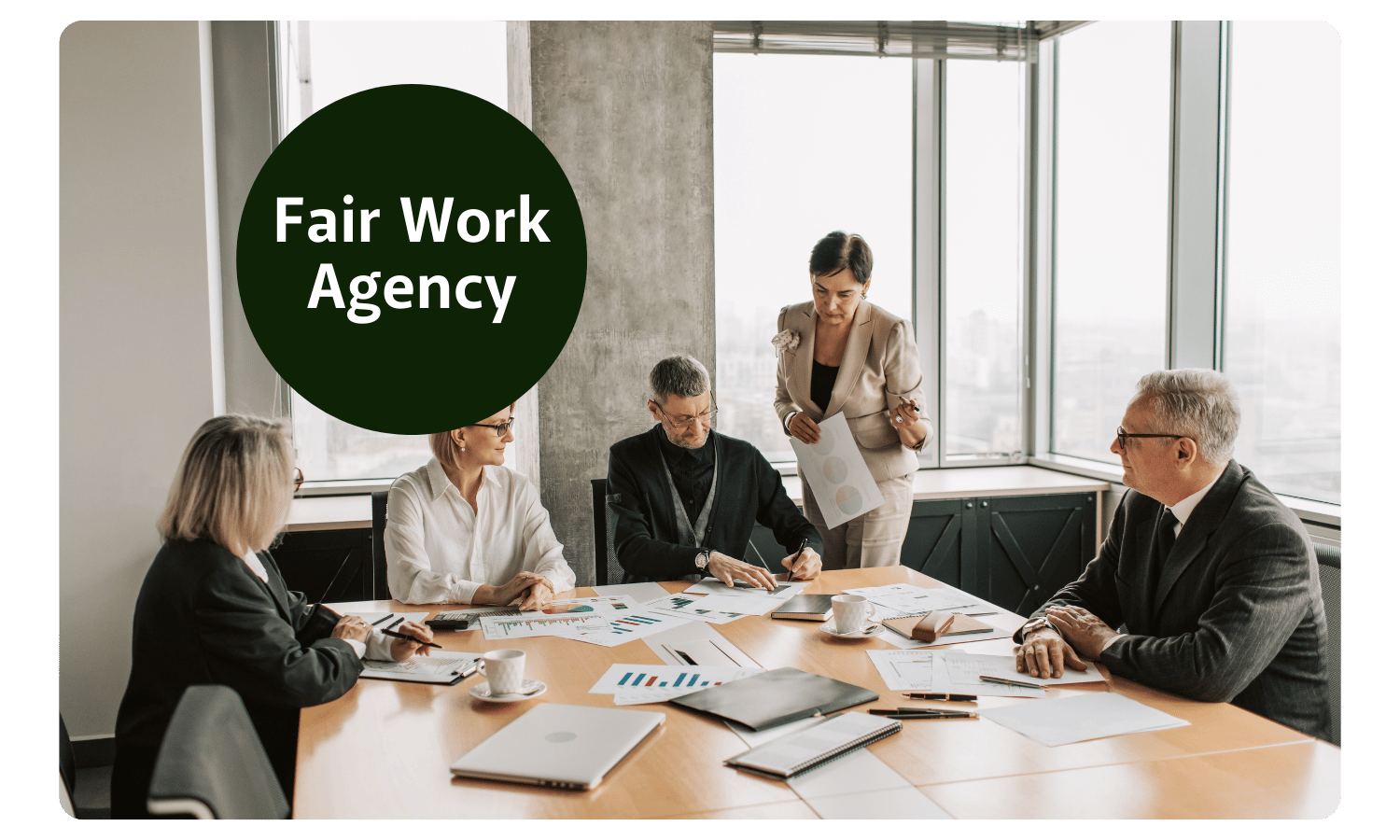 A group of professionally dressed people look at graphs and documentations on the table scattered between them. A woman holds a pen and looks down at a document as one of the men is writing on to it. It's a corporate office with large windows out to the city. In the top left of the image is 'Fair Work Agency' written