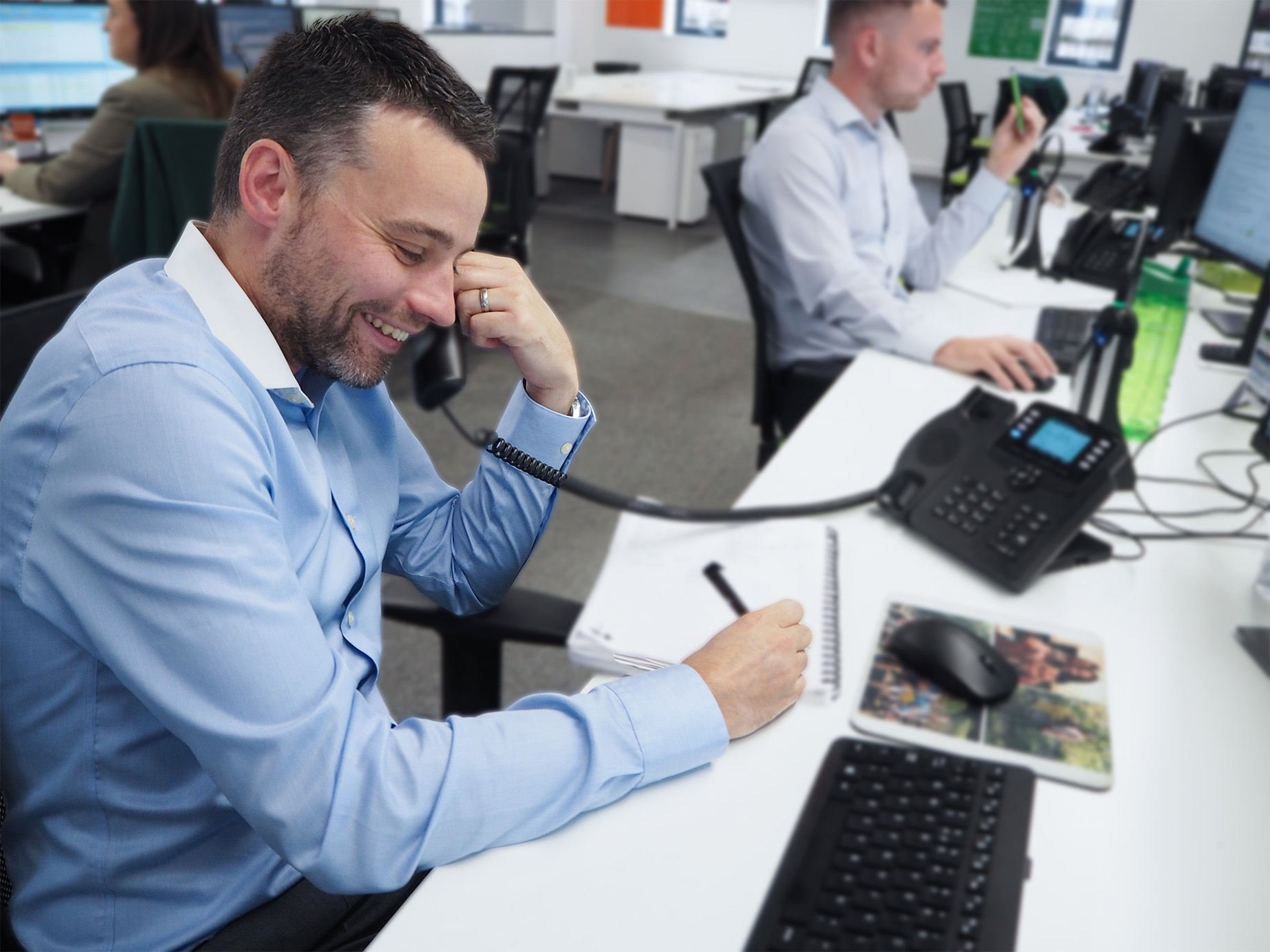 Mitchell smiles as he takes notes on a notepad, he's holding a phone in his hand speaking to a recruitment client. The Open-plan Prime appointments office is in the background.